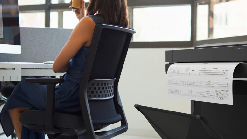 Lady sitting behind her desk looking at the HP app with a HP DesignJet printer in the background